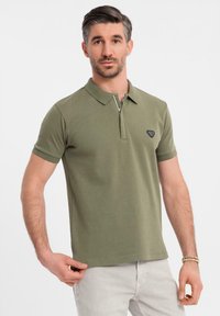 Olivově zelená polo košile vyrobená z jemné bavlny, s límečkem na zip a černou nášivkou s logem na levé části hrudi, s přiléhavými krátkými rukávy.