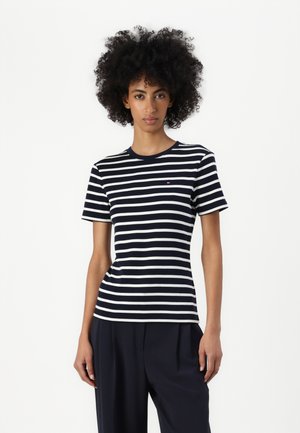 Žena s kovrčavom kosom, odjevena u tamnoplavu i bijelu kratku majicu s horizontalnim prugama i tamne široke hlače, stoji na bijeloj pozadini.