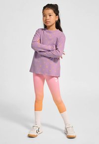 Paarse shirt met lange mouwen en geel driehoekpatroon, gecombineerd met roze ombre leggings en witte kniehoge sokken. Witte sneakers met bruine accenten.
