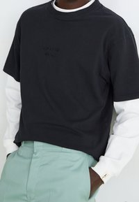 Zwart T-shirt met korte mouwen en een geborduurd logo, gedragen over een wit shirt met lange mouwen, gecombineerd met muntgroene broek.