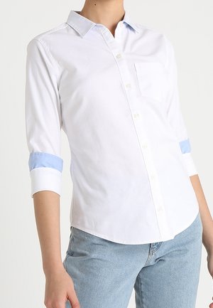 Hvid langærmet skjorte med knapper og lyseblå inderærmer, båret med lyseblå jeans på en person mod en ensfarvet baggrund.