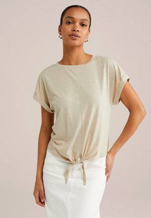 Beige kortærmet top med rund halsudskæring og knyttet frontdetalje, lavet af blødt stof, parret med en hvid nederdel.