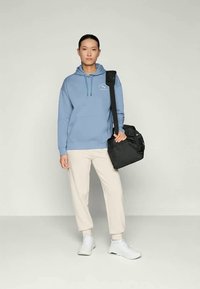 Felpa con cappuccio azzurro chiaro con tasca frontale, abbinata a pantaloni crema e sneakers bianche. Borsa a spalla nera con tracolla. Sfondo neutro.