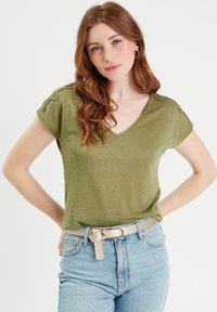 T-shirt verde oliva a maniche corte con scollatura a V e dettagli in pizzo sulle spalle, abbinato a jeans azzurri a vita alta e una cintura intrecciata.