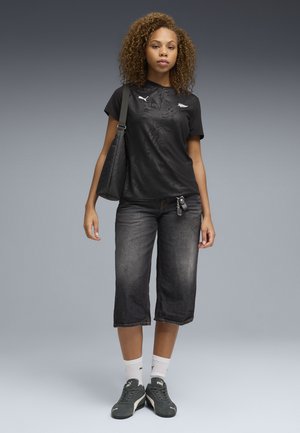 Jonge vrouw met krullend haar die een zwart Puma T-shirt draagt, verweerde zwarte cropped jeans, grijze Puma sneakers, witte sokken en een zwarte schoudertas bij zich heeft.