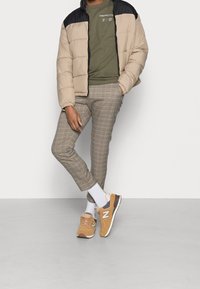 Chaqueta acolchada con cuerpo color beige y hombros negros, camiseta oliva, pantalones de cuadros y zapatillas New Balance marrones con suelas blancas.