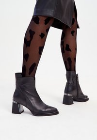 Zwarte leren enkelboots met een vierkante neus, geribbelde accenten en een stevige hak met een metallic detail. Draagt met met een patroonbedrukte panty.