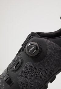 Černá cyklistická bota má pletený svršek s texturovanými akcenty, systém zapínání Boa a odolnou podešev s trakčními hřebeny.