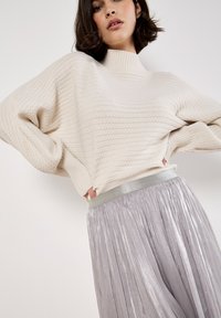 Suéter de crema grueso con textura acanalada y cuello alto, acompañado de una falda plateada plisada con una cinturilla metálica.