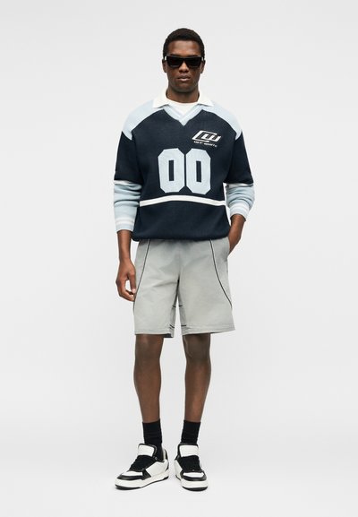 Άνδρας που φοράει μαύρα γυαλιά ηλίου, μια μπλε ναυτική και ανοιχτό μπλε αθλητική φανέλα με μακριά μανίκια και αριθμό 00, γκρι σορτς, μαύρες κάλτσες και μαύρα-άσπρα αθλητικά παπούτσια.