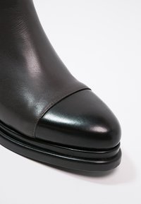 Bottine en cuir noir avec un bout arrondi, une finition brillante et une texture lisse, agrémentée d'un accent mat contrastant près du bout.