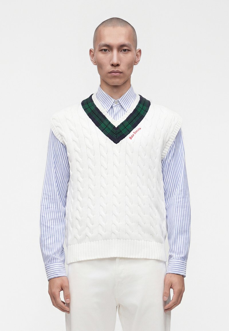 Homme portant un pull sans manches en laine torsadée blanc avec un col vert et bleu marine par-dessus une chemise rayée bleue à boutons, debout devant un fond blanc.