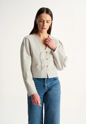 Kobieta o prostych brązowych włosach, ubrana w jasnoszarą koszulę w kratę zapinaną na guziki z bufkami oraz niebieskie dżinsy, patrzy w dół z ręką przy szyi.