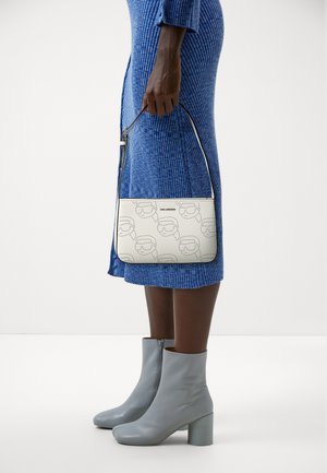 Borsa a spalla bianca in pelle con motivo impresso. Presenta una tracolla sottile e una chiusura con zip. Indossata con un vestito blu a coste e stivaletti grigi alla caviglia.