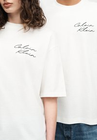 Δύο άτομα φορούν λευκά t-shirt Calvin Klein με μαύρο λογότυπο σε στυλ γραφής στο στήθος, το ένα με σγουρά μαλλιά και το άλλο με μπλε τζιν.
