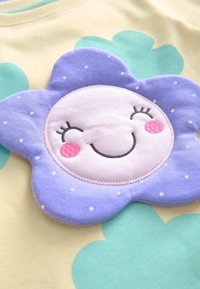 Chemise à manches courtes en tissu jaune clair avec une grande application en forme de fleur violette présentant un visage souriant aux joues roses et des pois blancs.