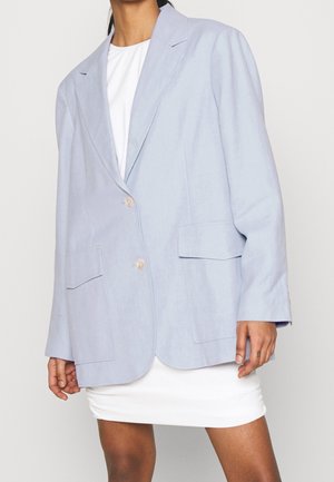 Mujer con un blazer azul claro de gran tamaño sobre un vestido blanco, de pie frente a un fondo liso gris claro.