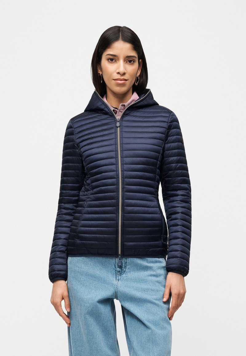 Γυναίκα που φοράει μπλε σκούρο καπιτονέ μπουφάν με κουκούλα και ανοιχτό μπλε τζιν, όρθια μπροστά σε λευκό απλό φόντο.