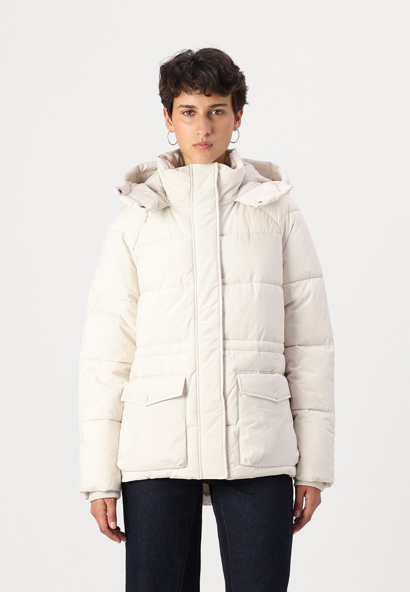 Doudoune beige clair avec col haut, fermeture éclair, capuche réglable et deux poches plaquées à l'avant. Texture lisse et matelassée.