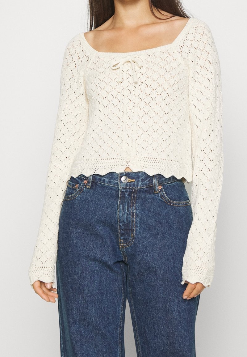 Pull en crochet blanc cassé, avec un décolleté carré, un devant noué et un ourlet festonné. Associé à un jean bleu taille haute.