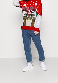 Person som bär en röd och vit jultröja med en ren-grafik, blå uppvikta jeans och vita sneakers, stående framför en enfärgad bakgrund.