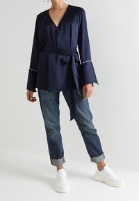 Top con envoltura en tejido suave con mangas acampanadas largas, acentos de ribete blanco y un detalle de lazo en la cintura, combinado con jeans de puños doblados y zapatillas.