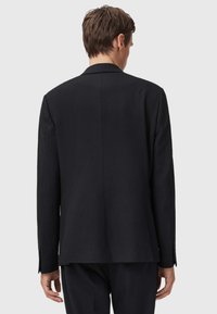 Uomo in piedi con le spalle alla macchina fotografica, che indossa un blazer nero aderente e pantaloni abbinati, su uno sfondo chiaro e semplice.