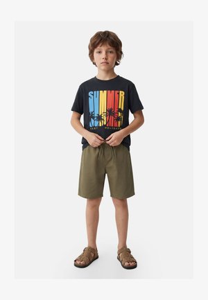 Niño joven con camiseta negra con la palabra "SUMMER" y diseño de palmera, pantalones cortos verde oliva y sandalias marrones, de pie con las manos cerca de la cintura.