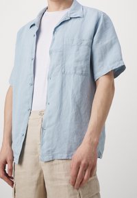 Camicia button-up a manica corta di colore azzurro chiaro, in una vestibilità rilassata, realizzata in un tessuto testurizzato, con taschino sul lato sinistro del petto e una finitura morbida.