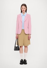 Rosa Blazer, hellblaues Hemd, beigefarbene, plissierte Shorts, schwarze Handtasche, weiße Socken und klobige schwarze Schuhe mit einer Schnallendetail.