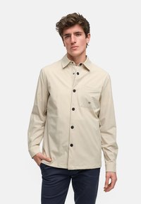 Beige skjorte med knapper, svarte knapper, krage og brystlomme. Ensfarget tekstur med en subtil logo på lommen. Lange ermer.