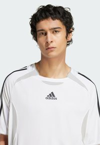 Balta sportinė marškinė su trumpomis rankovėmis, turinti juodas šonines detales, apvalų kaklą ir juodą "Adidas" logotipą krūtinėje.