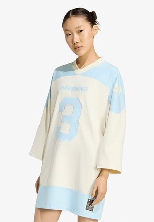 Joven mujer con vestido camisola de Adidas de gran tamaño en beige y azul claro con el número 3, de pie frente a un fondo blanco.