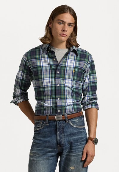 Rutet skjorte i grønn, blå og hvit, lagdelt over en grå t-skjorte. Bært med falmede blå jeans og et brunt belte.
