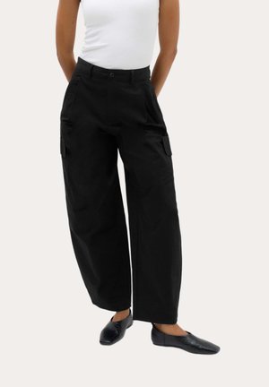 Donna che indossa pantaloni cargo neri a gamba larga, scarpe nere basse e una canotta bianca senza maniche, in piedi con le mani dietro la schiena.