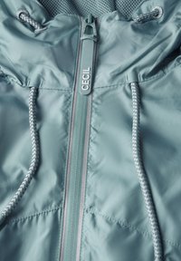 Veste bleu clair avec fermeture éclair sur le devant portant le nom de la marque, capuche avec cordon de serrage et accents texturés, et tissu lisse et brillant.