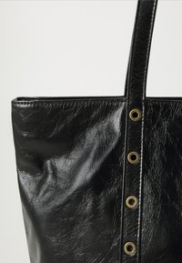 Svart läderhandväska med vertikal rem som har fyra mässingsöglor mot en plain vit bakgrund.