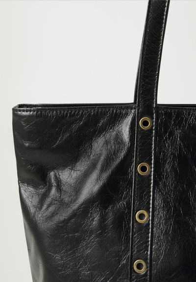 Sac fourre-tout en cuir noir avec une sangle verticale comportant quatre œillets en laiton sur fond blanc.