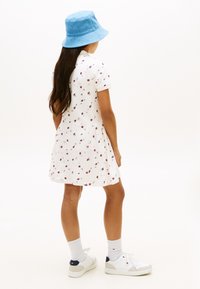 Fată purtând o rochie albă cu mâneci scurte și imprimeu floral, pălărie tip găleată albastră, șosete albe și adidași albi, stând cu fața spre spate pe un fundal simplu.