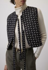 Crni prsluk s uzorkom i vezanjem, s bijelim apstraktnim dizajnom. Nosivši ispod kremasti džemper s rolkom, uparen s sivim hlačama.