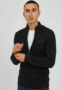 Black ribbed zip-up cardigan with a high collar, textured pattern, and a small logo detail at the hem. Worn over a white shirt.