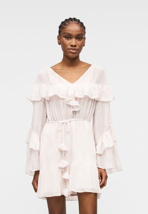 Junge Frau mit geflochtenem Haar, die ein weißes, langärmliges Kleid mit Rüschen und einem Kordelzug in der Taille trägt, vor einem schlichten Hintergrund stehend.