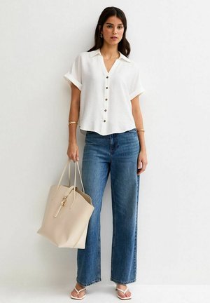 Femme portant un chemisier blanc à manches courtes avec boutons, un jean droit bleu, des sandales blanches, tenant un sac fourre-tout beige, debout contre un mur blanc.