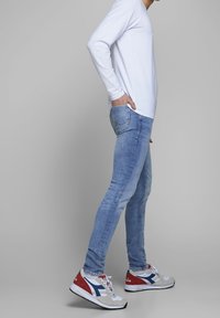 Camisa blanca de manga larga, vaqueros ajustados de color azul claro y zapatillas multicolores con acentos en rojo, azul y gris. Diseño limpio y moderno.