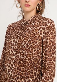 Blusa animalier in marrone e crema. Presenta un colletto, una patta con bottoni e una tasca sul petto. Tessuto morbido e strutturato con vestibilità ampia.