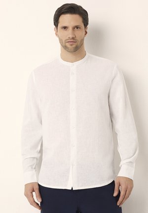 Camisa de lino blanca con cuello mao, cierre de botones en la parte delantera, mangas largas y una superficie ligeramente texturizada, combinada con pantalones oscuros.