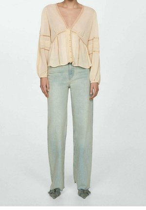 Femme portant un chemisier beige clair à manches longues avec des boutons, associé à un jean bleu clair à jambes larges et des chaussures vertes pointues.