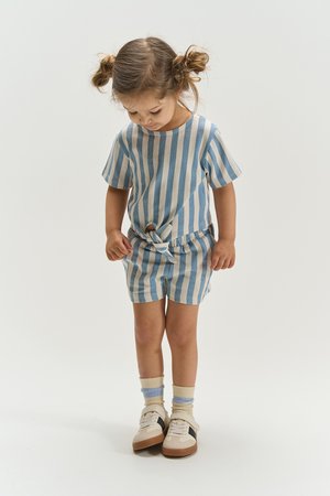 Jong meisje met bruin haar in twee knotjes, gekleed in een blauw-beige gestreepte korte mouwen top en korte broek, kijkt naar beneden en staat tegen een witte achtergrond.