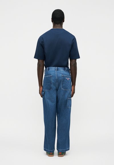 Μαύρος άνδρας που στέκεται με την πλάτη, φοράει μπλε ναυτικό κοντομάνικο πουκάμισο, φαρδιά μπλε τζιν με τσέπες με επιδιορθώσεις και πολύχρωμα παπούτσια, σε απλό φόντο.