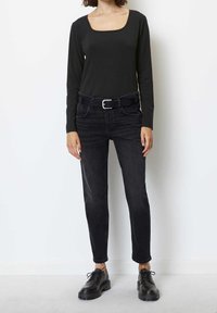 Kvinna klädd i svart långärmad topp med rundad halsringning, svart skärp, svarta åtsittande jeans och svarta snörade klumpiga skor stående mot en vit vägg.